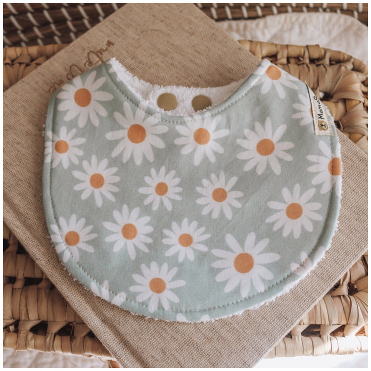Handmade baby bib with white daisy flowers printed on a pale turquoise blue background, backed with white terry towelling, styled on natural woven fabric.