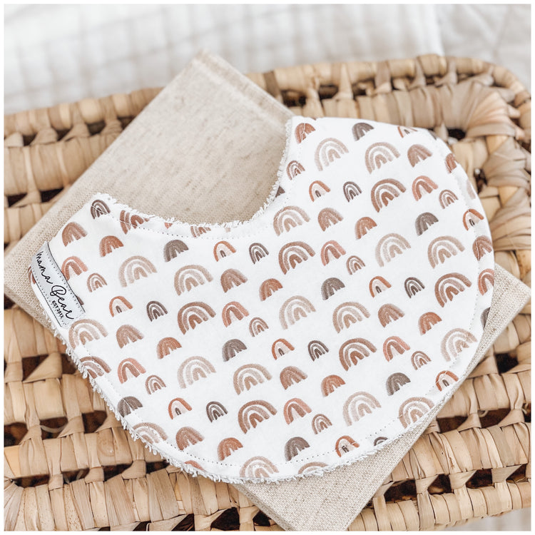 Neutral baby bib with warm-toned rainbow print in rust, beige, and brown shades, styled on a linen cloth over a woven basket.