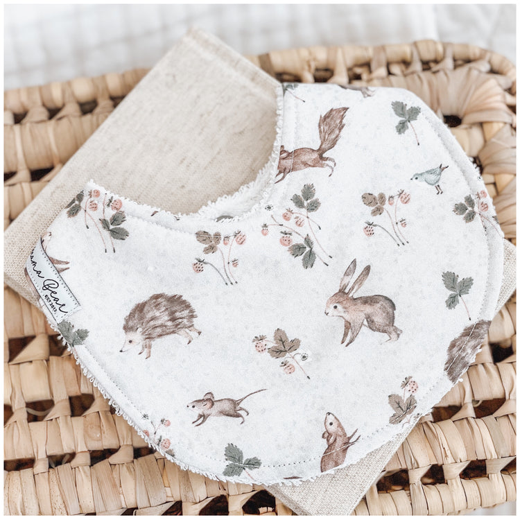 Handmade baby bib with an illustrated woodland animal print, including rabbits, hedgehogs, mice, squirrels, birds, and strawberry vines on a soft cream background. Bib is laid on a beige cloth over a woven basket.