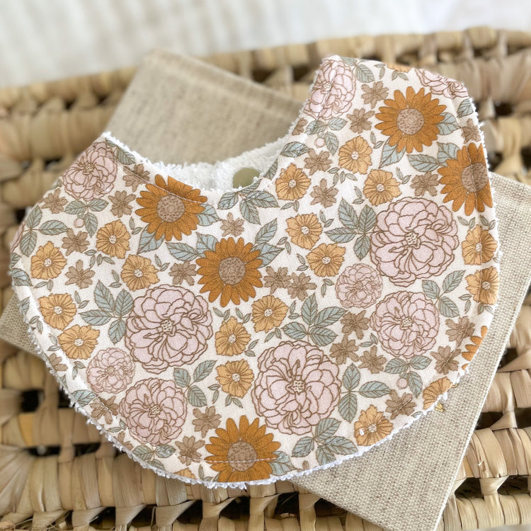 Retro floral baby bib with mustard, blush, and beige flowers on an off-white background. Bib is laid flat on a woven mat with a neutral linen cloth underneath.