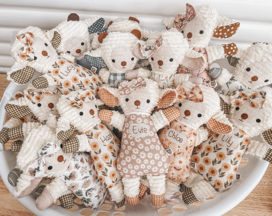 Unique baby gift ideas. Handmade lamb toy personalised with babies name. Basket shows a bunch of personalised lambs in different pastel fabrics. The girl lambs have a mini hair bow and the boy lambs have little horns next to their ears. 