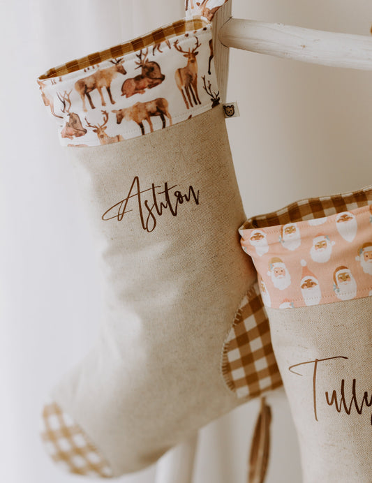 A brown neutral Christmas stocking with reindeer pattern, personalized with the name 'Ashton' in script font, hanging against a white wall.