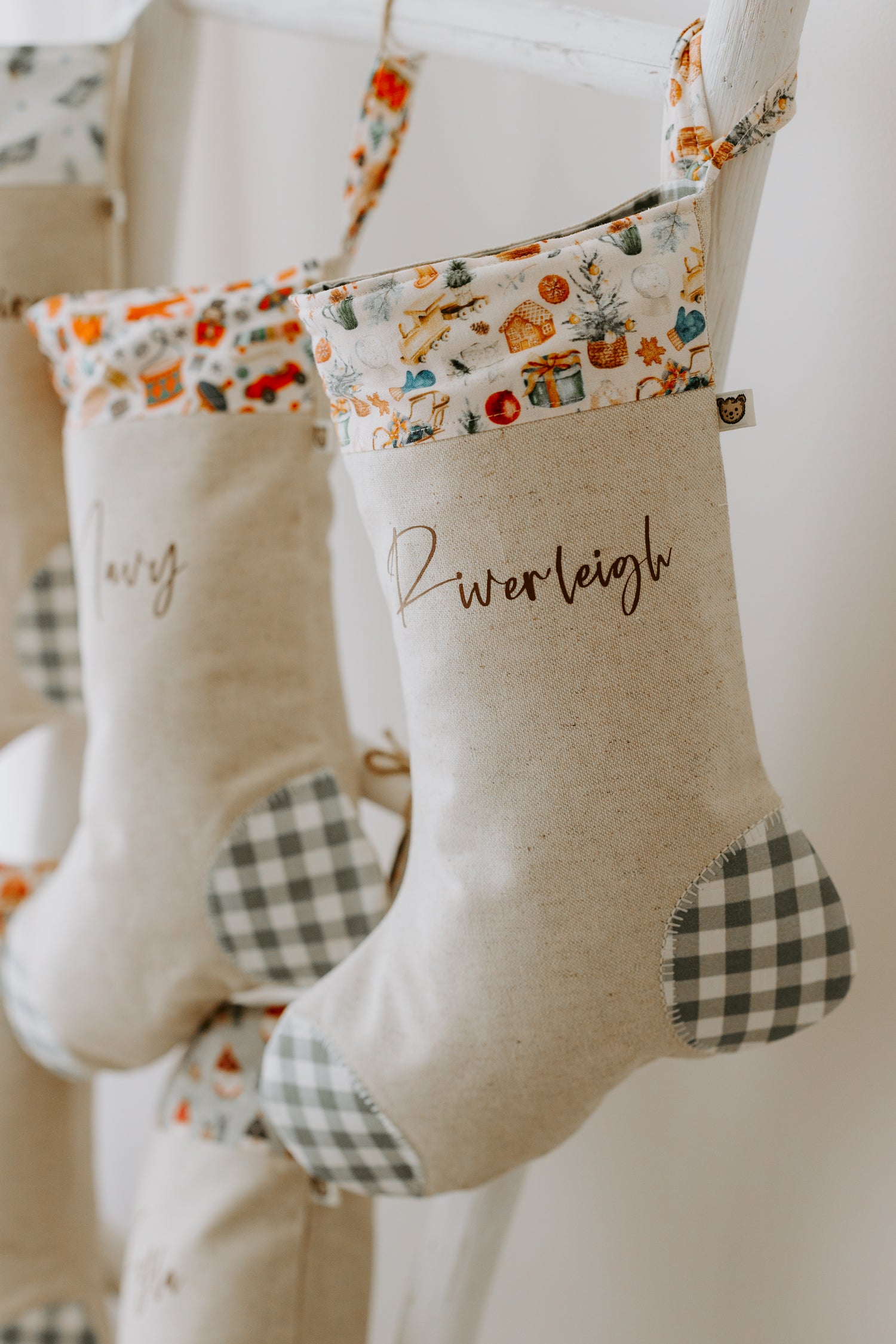 Personalised Christmas stocking handmade in beige fabric with green gingham heel and toe and festive Christmas cuff featuring toys, baubles and gifts, customised with name Riverleigh.