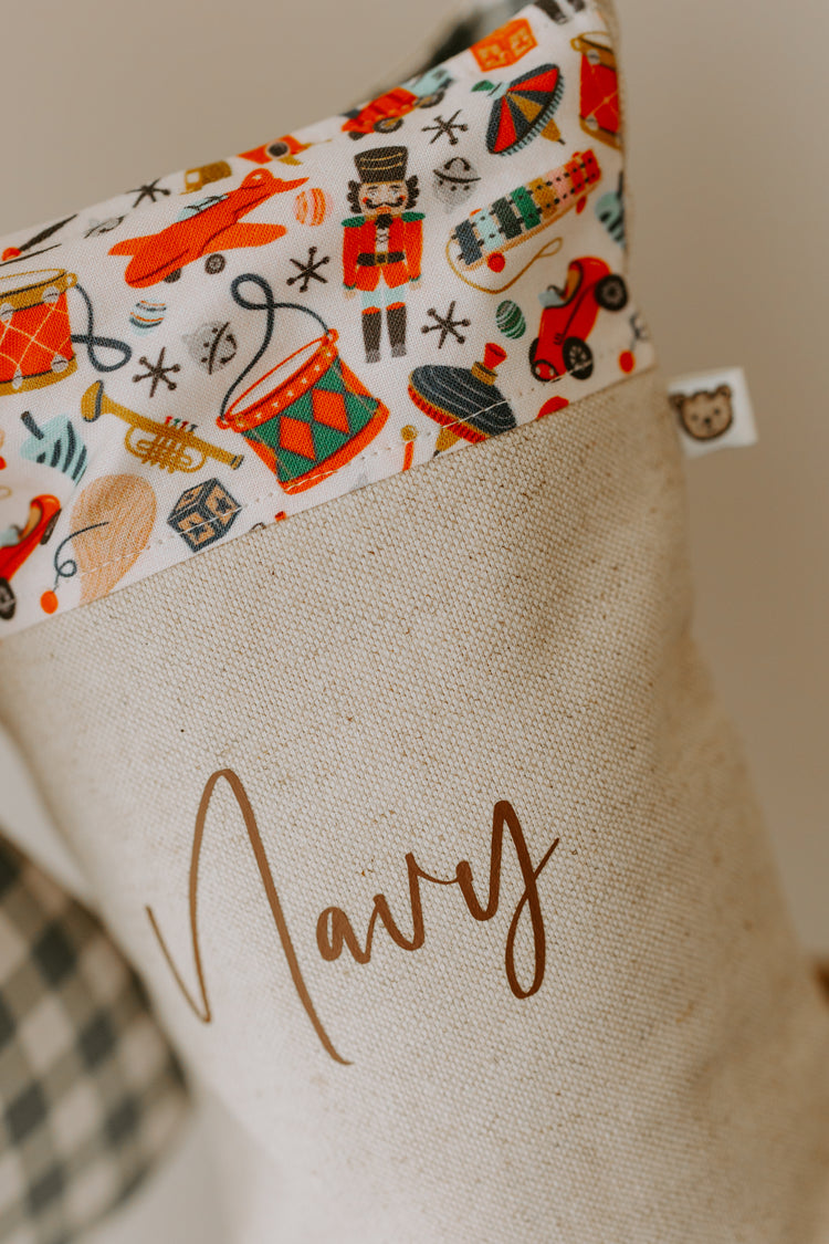 Close-up of a custom linen Christmas stocking with the name “Navy.” The stocking’s cuff is decorated in a retro Christmas toy pattern, showcasing bright red, green, and gold nutcrackers, drums, and holiday figurines. Green gingham fabric details at the heel and toe add a farmhouse-style touch, making this stocking a unique keepsake for children and families.