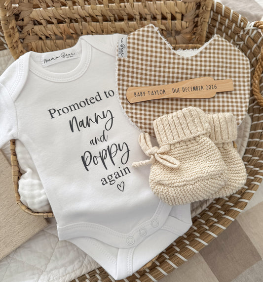 A baby nursery gift set arranged in a woven wicker basket, featuring a white bodysuit with 'Promoted to Nanny and Poppy again' text, a brown and cream gingham bib, and a pair of cream knit booties. A wooden pregnancy test that reads 'BABY TAYLOR - Due December 2016'.