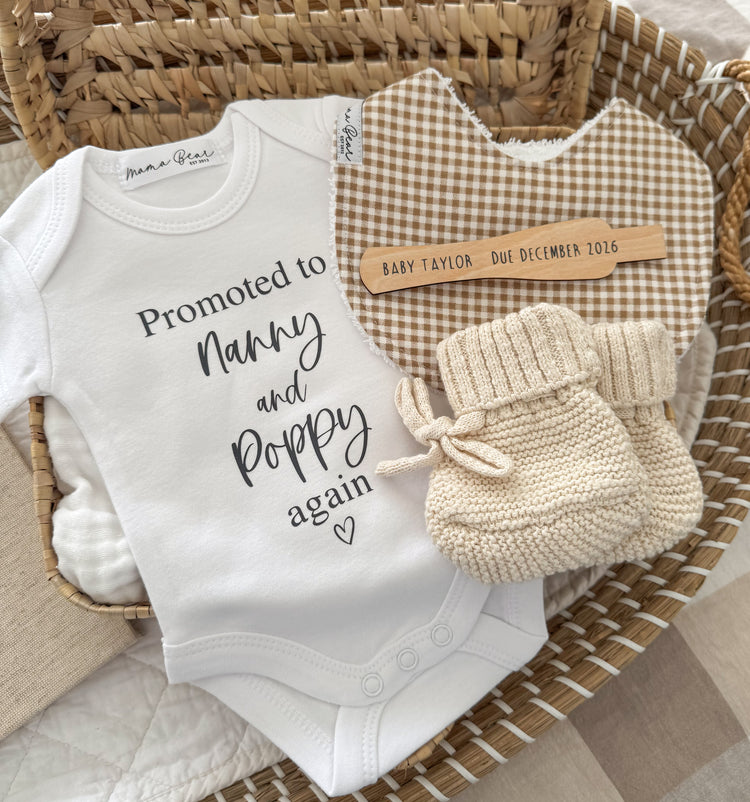 A baby nursery gift set arranged in a woven wicker basket, featuring a white bodysuit with 'Promoted to Nanny and Poppy again' text, a brown and cream gingham bib, and a pair of cream knit booties. A wooden pregnancy test that reads 'BABY TAYLOR - Due December 2016'.