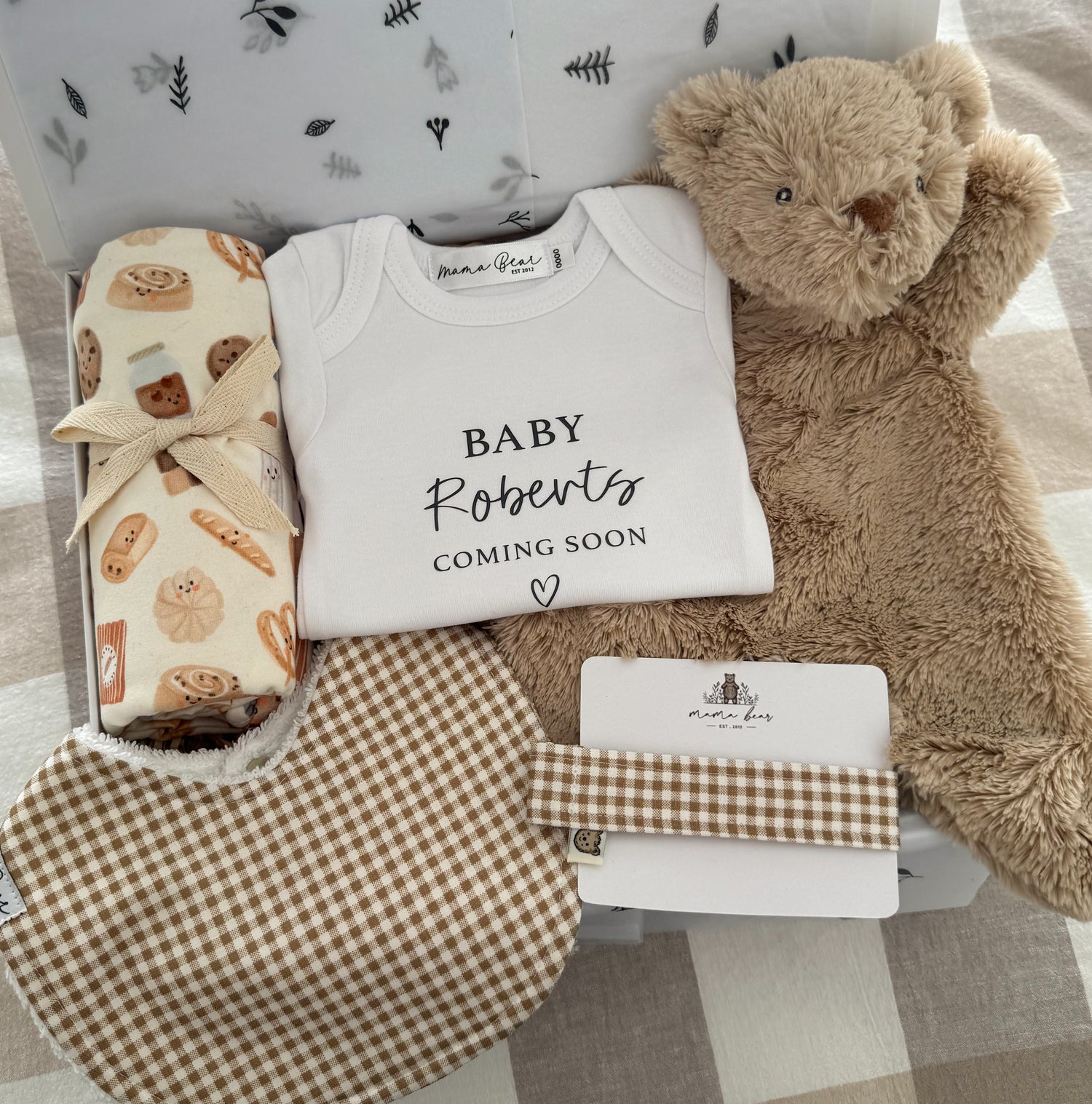 Baby shower gift set with personalsied baby onesie 'Baby Roberts coming soon', soft brown teddy bear comforter, brown gingham check baby bib & matching dummy clip and a cream swaddle with cute illustrations of bakery foods such as danishes and bread. Packaged inside a white gift box.