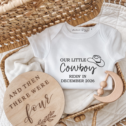 Western cowboy inspired baby onesie 'Our little cowboy ridin' in December 2026'. Onesie is white with black text also a small tilted cowboy hat illustration on onesie.