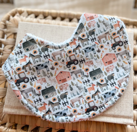 Handmade baby bib with a farmyard pattern including animals, tractors, barns, and sunflowers in muted tones; backed with absorbent terry towelling and suitable for newborns and teething babies.