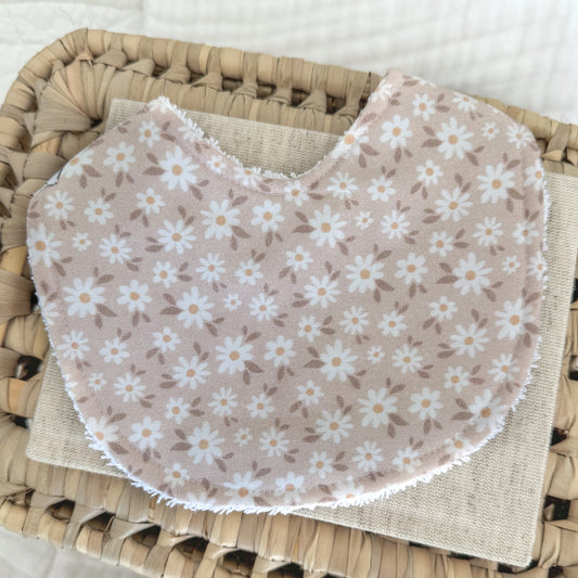 Contoured baby bib with a delicate white daisy print and golden-yellow centres on a soft blush-taupe cotton background, handmade and perfect for gifting a newborn.