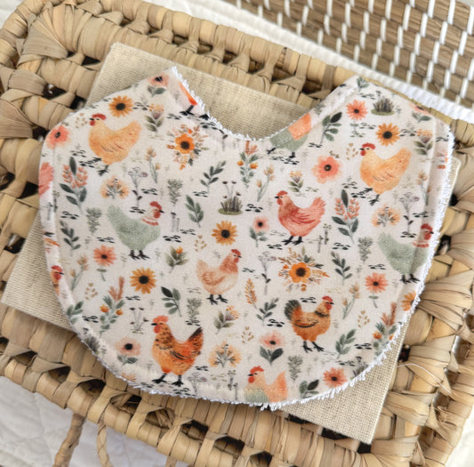 Baby bib: Background beige, illustrations of hens and wildflowers scattered. Main colours are yellows and sage green.