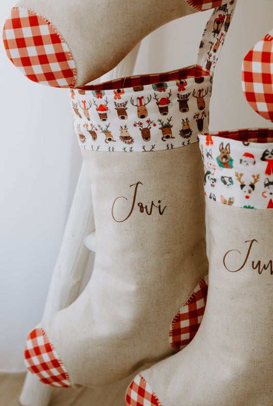 Handmade personalised Christmas stocking in natural linen, featuring a festive cuff with playful reindeer illustrations wearing Christmas hats and antlers, finished with bold red gingham fabric on the heel and toe. The stocking is customised with the name “Jovi” in brown script font, creating a unique keepsake gift for children or families to hang by the fireplace.