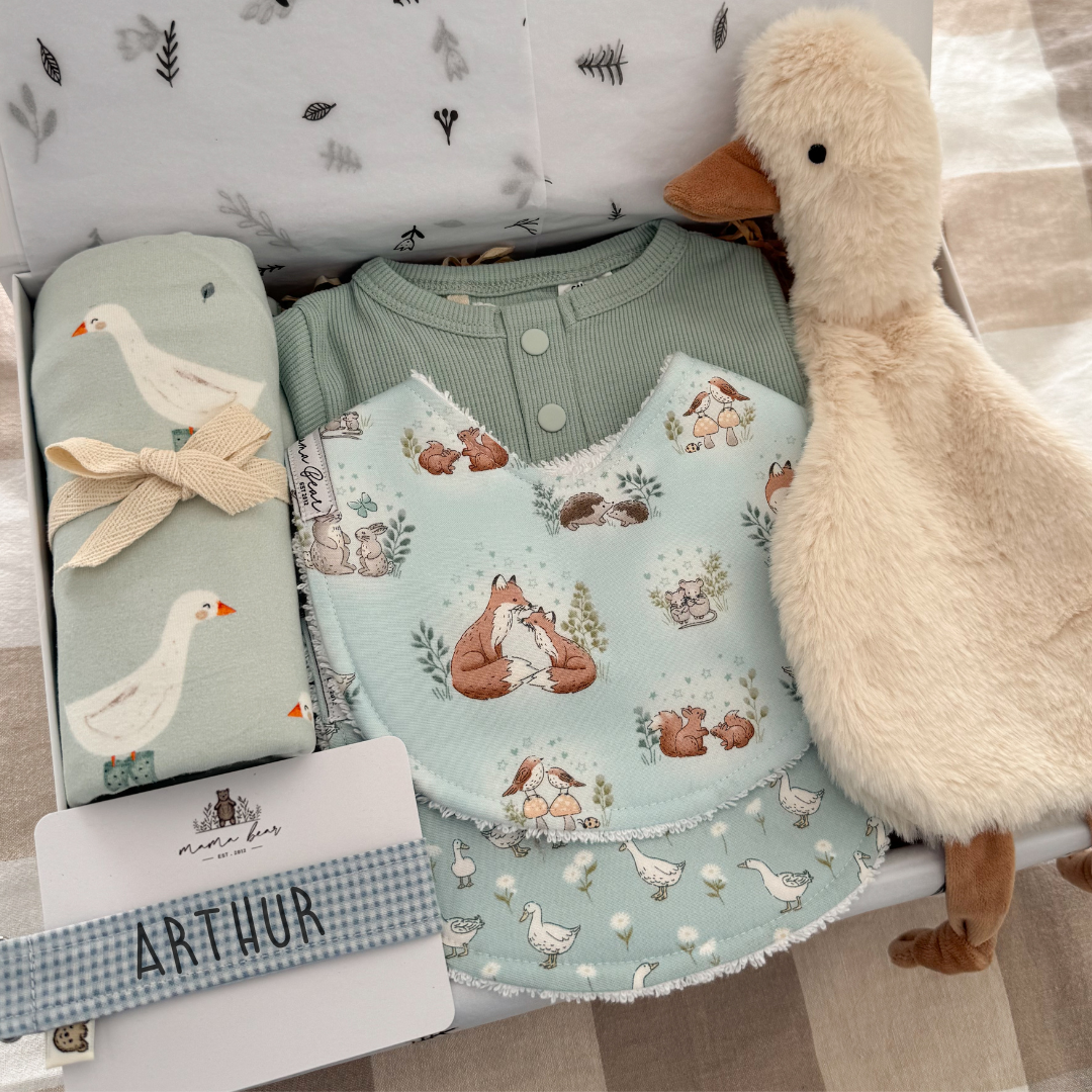 A baby boy gift set arranged in a white magnetic box, featuring a sage green ribbed henley bodysuit, a light blue bib with woodland animal prints, a matching rolled swaddle with white geese and a pale blue background,  a blue gingham personalised dummy clip, and a plush goose toy. 