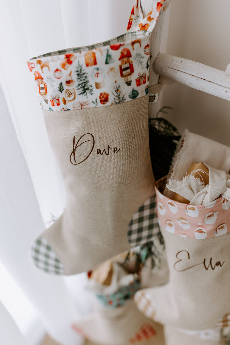 Personalised Christmas stocking for kids and adults with the name Dave in script font. Handmade in Australia from natural linen with green gingham heel and toe accents and a festive cuff featuring nutcrackers, Christmas trees, holly, and gingerbread. Stylish farmhouse Christmas décor idea, perfect as a unique adult holiday gift or men’s Christmas stocking to hang by the fireplace or under the tree.
