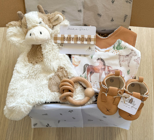 A newborn gift hamper with farm theme containing a plush cow toy, cotton bibs, a personalized dummy clip, a wooden ring rattle, and baby moccasins presented in a gift box.