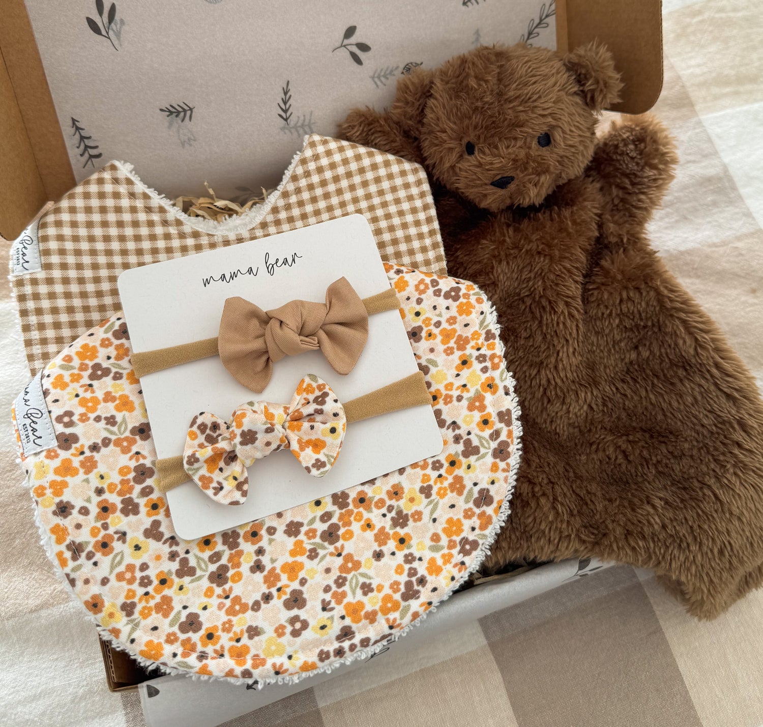 A baby gift set in a kraft box featuring a floral bib in orange, yellow and brown tones, as well as a coordinating bib in mini brown gingham check, a brown teddy bear plush toy, and 2 matching bow hairbows in the same fabrics.