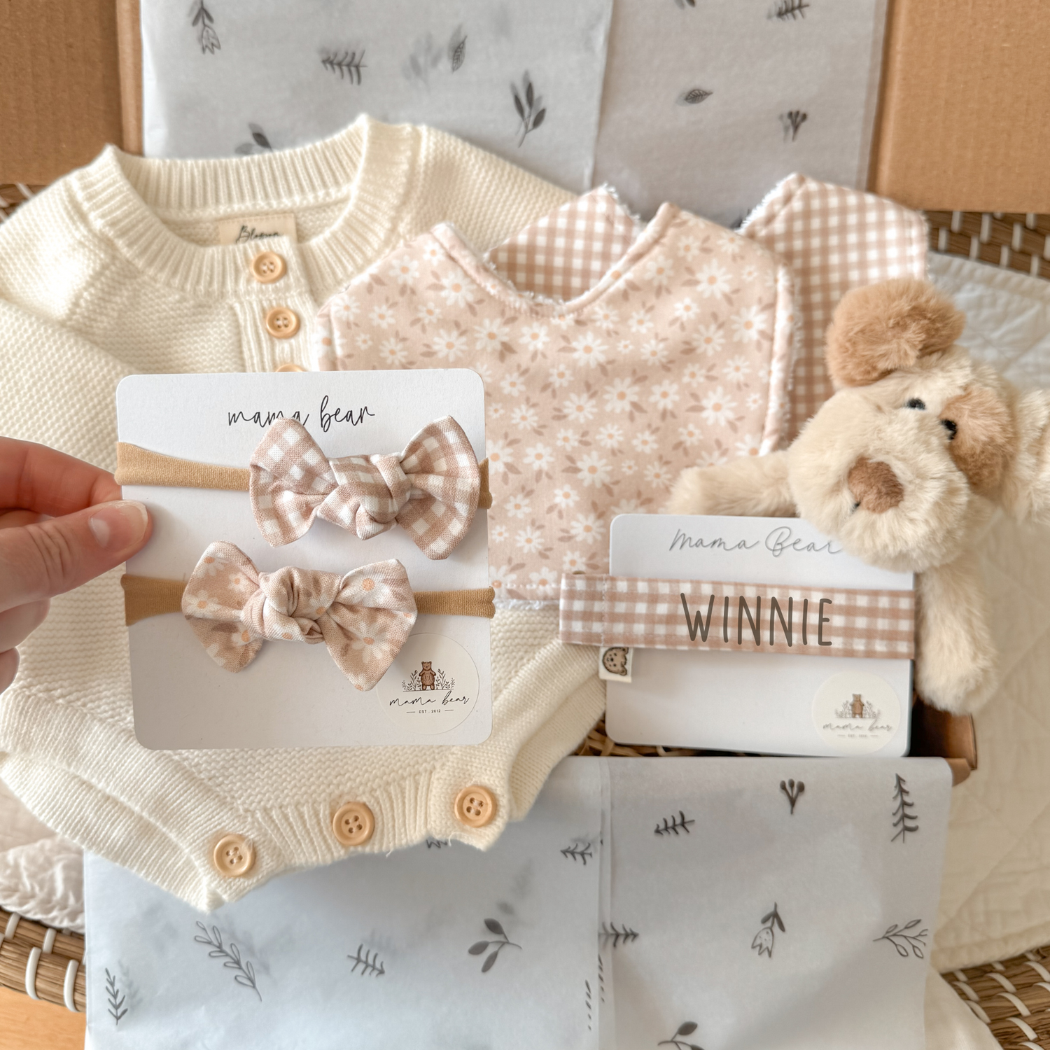 A beautifully arranged baby gift box featuring soft neutral tones. The box includes a cream knitted cardigan with wooden buttons, a floral bib, and two matching headbands with gingham and floral bows on a Mama Bear display card. A personalized gingham name headband with “Winnie” printed on it is included, along with a soft, plush puppy toy. The items are neatly arranged on decorative tissue paper, creating an elegant and thoughtful presentation.