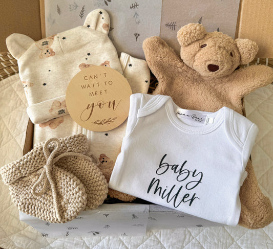 Gender-neutral baby announcement gift hamper featuring a soft beige teddy bear, personalised baby announcement onesie, wooden milestone disc, and coordinating baby essentials, all beautifully arranged in a gift box with neutral tones and natural textures, perfect for baby showers or special announcements. 