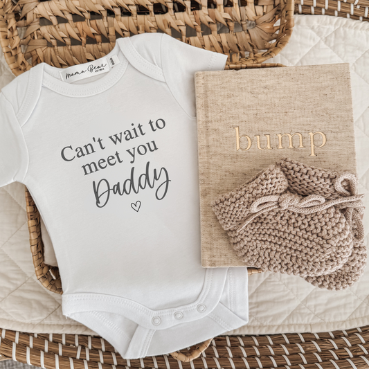 Pregnancy announcement , baby reveal to partner. White baby onesie that says in black text 'can't wait to meet you' (in times roman font), 'Daddy' (in script font) with a small dainty heart underneath. Photo props in flat lay image also include an oatmeal linen 'bump' pregnancy journal with a pair of beige booties. 