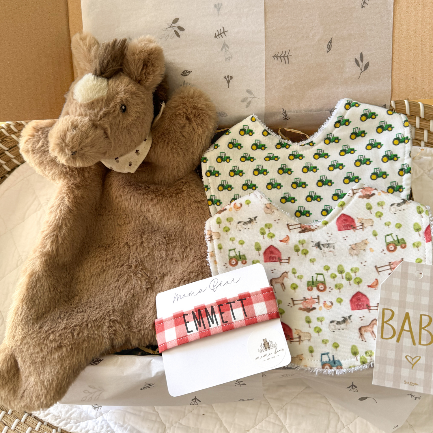 Unisex baby gift box featuring a farm animal themed bib, green tractor bib, a personalised dummy clip in red check & a brown baby horse toy comforter. The items are nestled in a gift box with shredded paper for a rustic, farm inspired presentation—perfect for baby showers or newborn gifts.