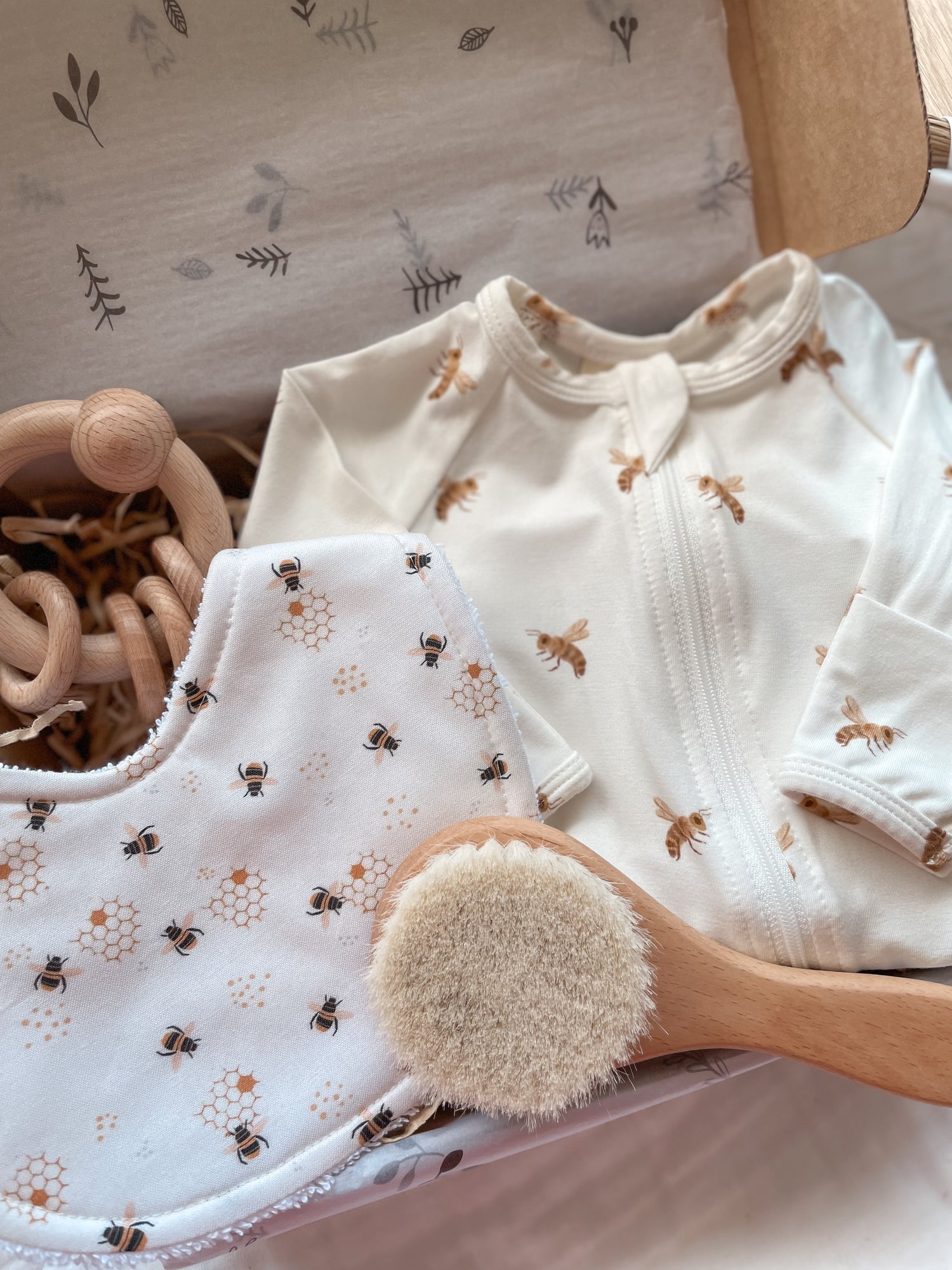 Gender neutral baby gift box featuring handmade bib and blossom & pear zipsuit with honey bee pattern on white background. Presented in a Kraft gift box filled with tissue paper and raffia. Also shown in the set is a wooden round baby hairbrush and a round wooden ring rattle.