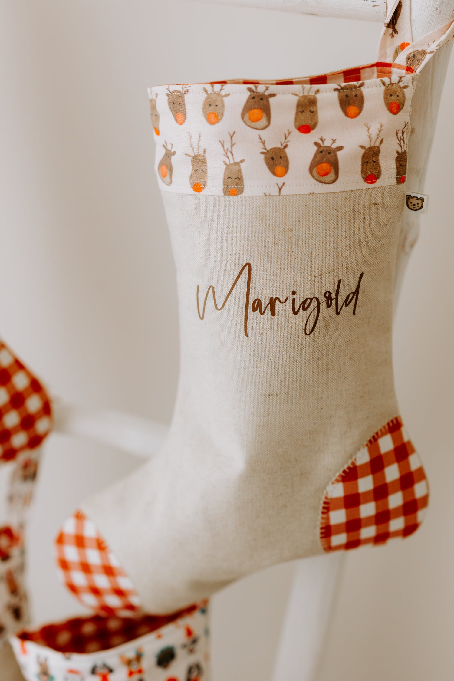 Handmade personalised Christmas stocking in beige fabric with red gingham heel and toe, featuring a reindeer print cuff, customised with name Marigold.