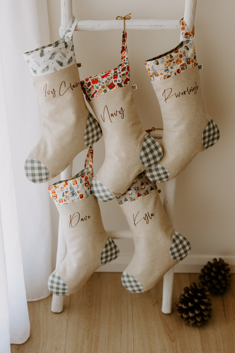 Set of five personalised Christmas stockings hanging on a rustic white ladder, handmade in beige linen with grey gingham heel and toe patches. Each stocking features a unique cuff design — pine branch print, vintage toy print, festive village, Santa faces, and Christmas trees — with names “Ivy Claire,” “Navy,” “Riverleigh,” “Dave,” and “Kyra” in script font. A warm and festive family display, perfect for boys, girls, and adults, adding personalised charm to holiday décor.