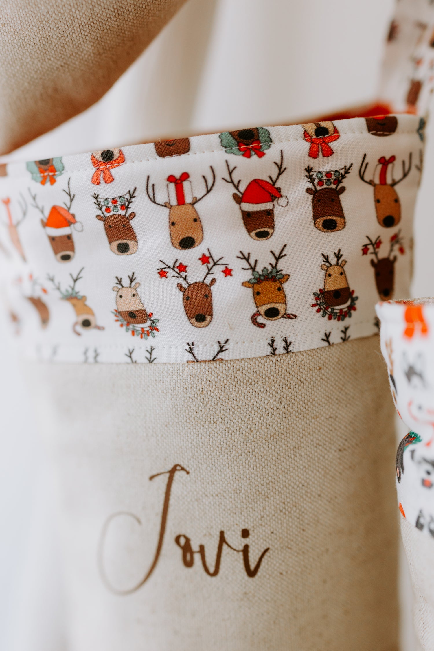 Close-up of a handmade Christmas stocking cuff featuring whimsical reindeer illustrations in festive hats, wreaths, and antlers, printed on white fabric. The stocking is personalised with the name “Jovi” in brown script on natural linen