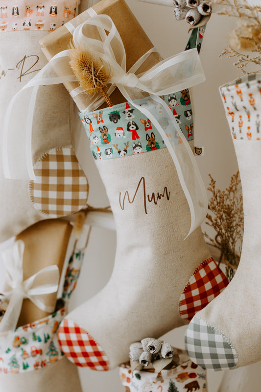 Handmade personalised Christmas stocking with “Mum” written in script font, featuring a cuff decorated with festive dog illustrations wearing Santa hats, scarves, and wreaths. The stocking has a natural linen body with red and green gingham patches and is styled with a wrapped gift and rustic holiday décor. Perfect pet-themed Christmas stocking for dog lovers and families in Australia.