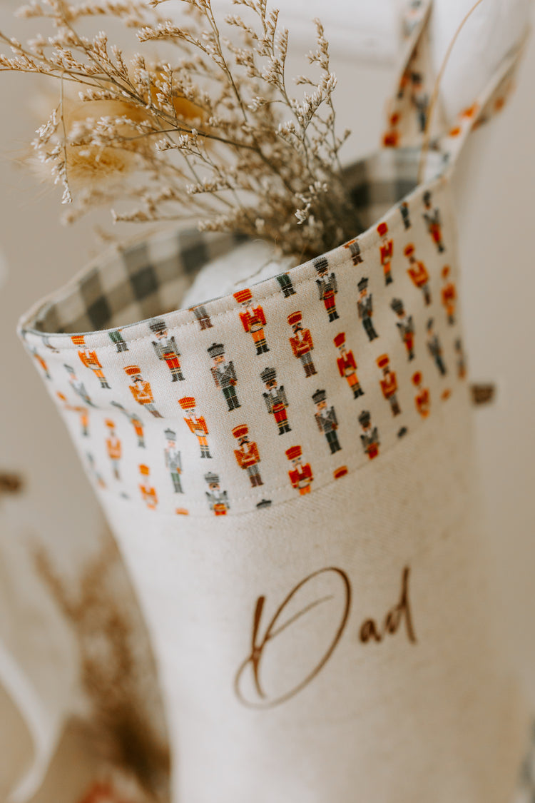 Close-up of handmade personalised Christmas stocking in beige fabric with nutcracker soldier print cuff and green gingham trim, customised with name Dad.