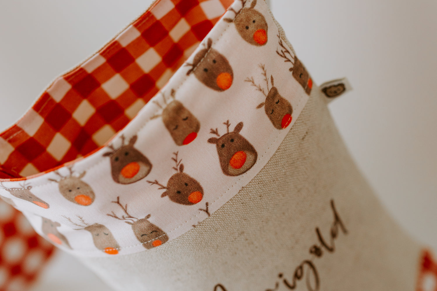 Christmas stocking handmade in Australia, close up of beige stocking, trim is illustrations of Rudolph the reindeer faces with a red nose, inside stocking trim is a red gingham. Personalised with name. 