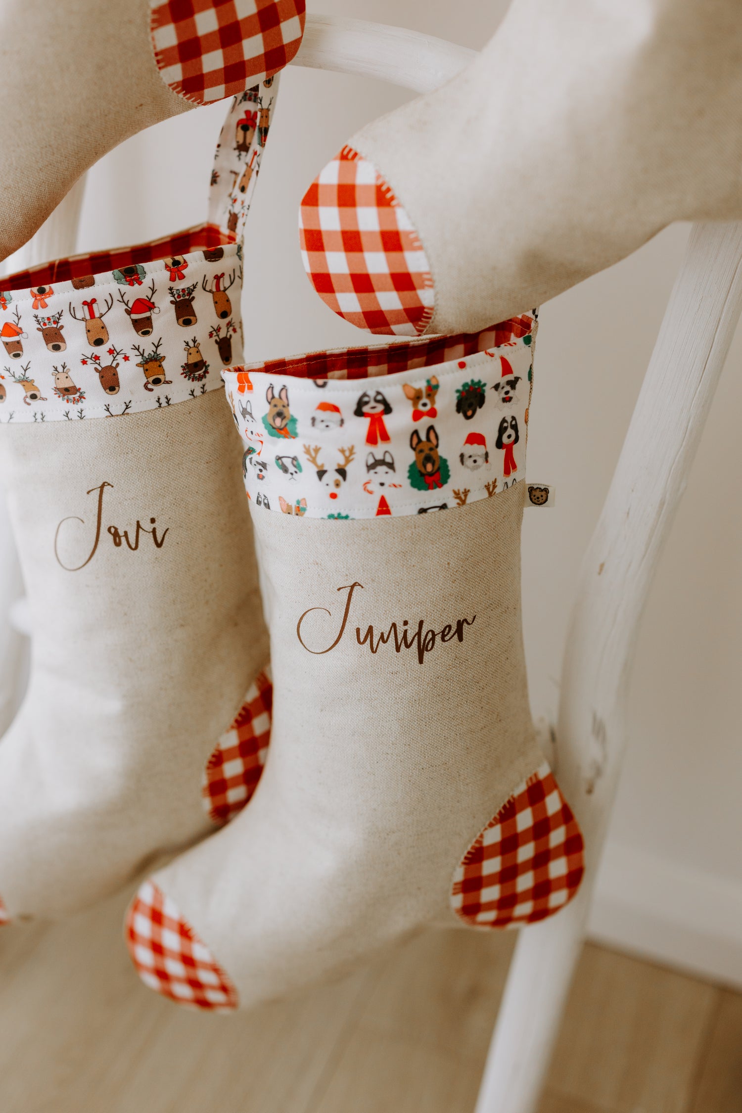 Two personalised linen Christmas stockings hanging on a white wooden ladder, each with bold red gingham heel and toe patches. The stocking on the left reads “Jovi” and features a cuff with playful reindeer faces wearing Santa hats and festive wreaths. The stocking on the right reads “Juniper” and has a cuff patterned with cheerful dog illustrations — including huskies, spaniels, and shepherds dressed in Christmas scarves, wreaths, and Santa hats. A charming handmade holiday stocking set, perfect for familie