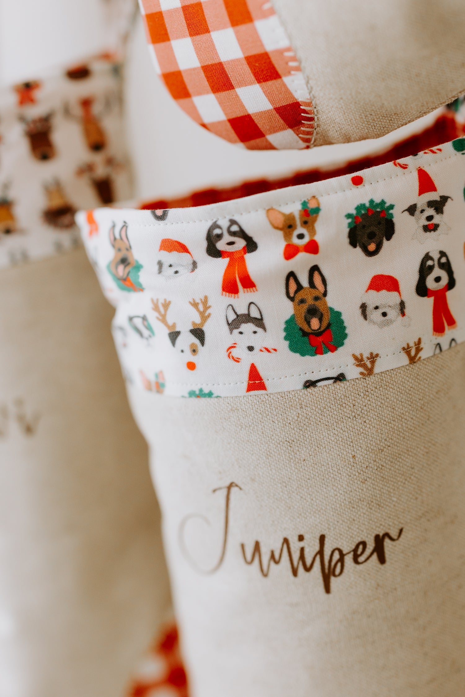 Close-up of a personalised Christmas stocking made from natural linen with red gingham heel patch, featuring the name “Juniper” in brown script. The cuff fabric is decorated with playful dog illustrations dressed for Christmas — including puppies wearing Santa hats, red scarves, reindeer antlers, and festive wreaths. A unique handmade Christmas stocking that celebrates pets, perfect for dog lovers or families who want to include their furry friends in holiday traditions.