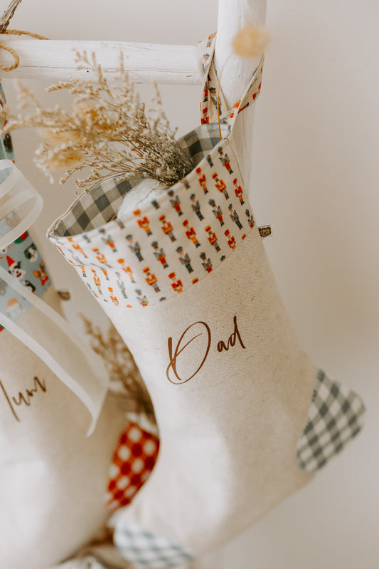 Handmade personalised Christmas stocking in beige fabric with green gingham heel and toe, featuring a nutcracker soldier print cuff, customised with name Dad.