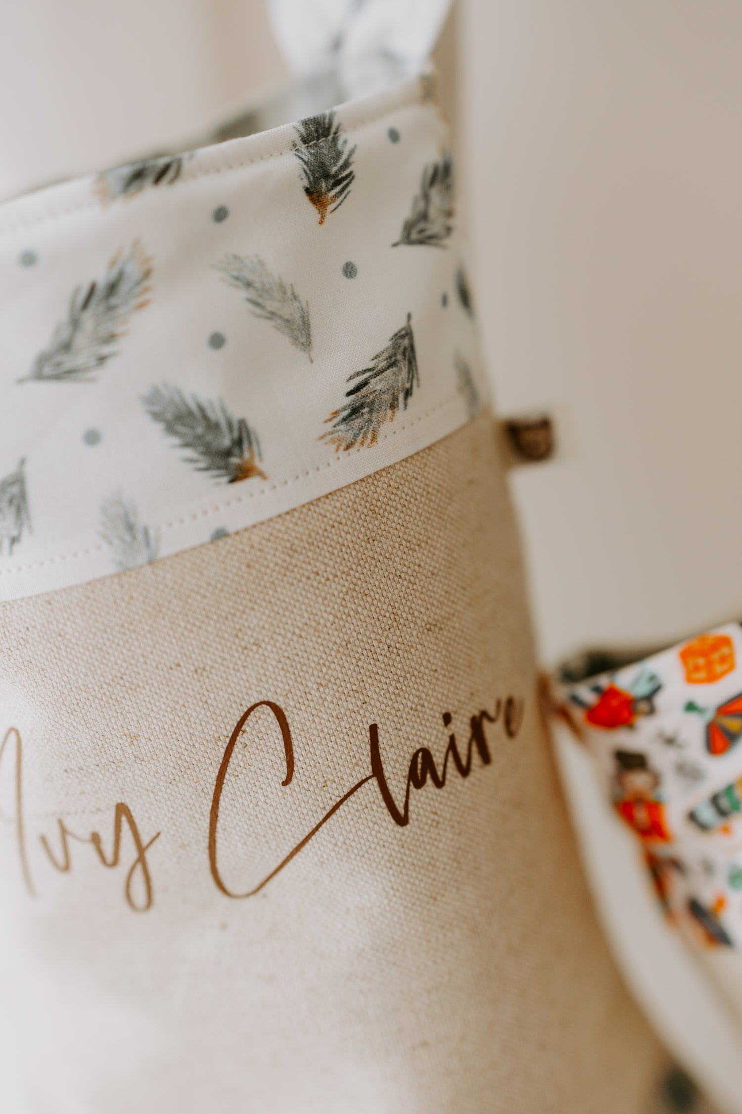 Close up of Christmas stocking, beige stocking with name Ivy Claire in brown script don’t. Trim of stocking is white with scattered pine leaf branches, making for a gender neutral stocking.