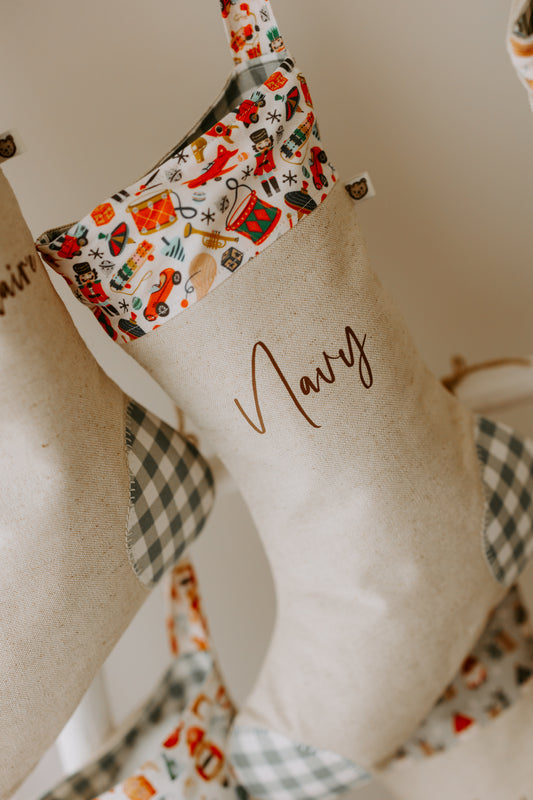 Personalised Christmas stocking in natural linen fabric with the name “Navy” in brown script. The cuff features a colourful vintage toy print with nutcrackers, toy drums, rocking horses, and Santa motifs, bringing a nostalgic festive feel. Finished with sage green gingham heel and toe patches, this handmade stocking combines rustic with playful holiday tradition.