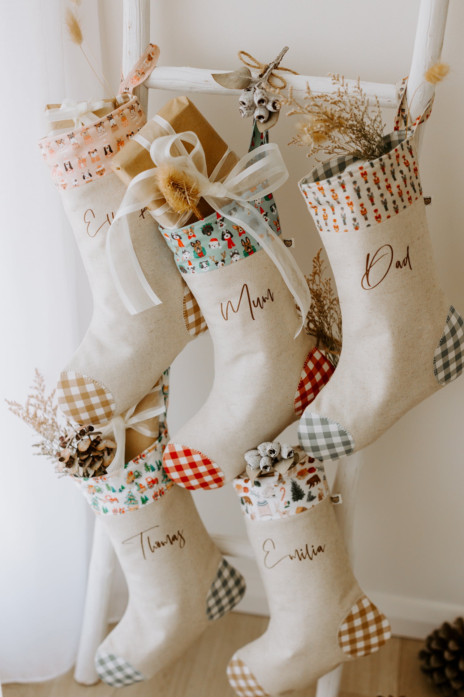 Set of handmade personalised Christmas stockings in beige fabric with gingham heels and assorted festive cuffs, customised with names Emilia, Mum, Dad and Thomas, filled with rustic holiday décor and gifts.