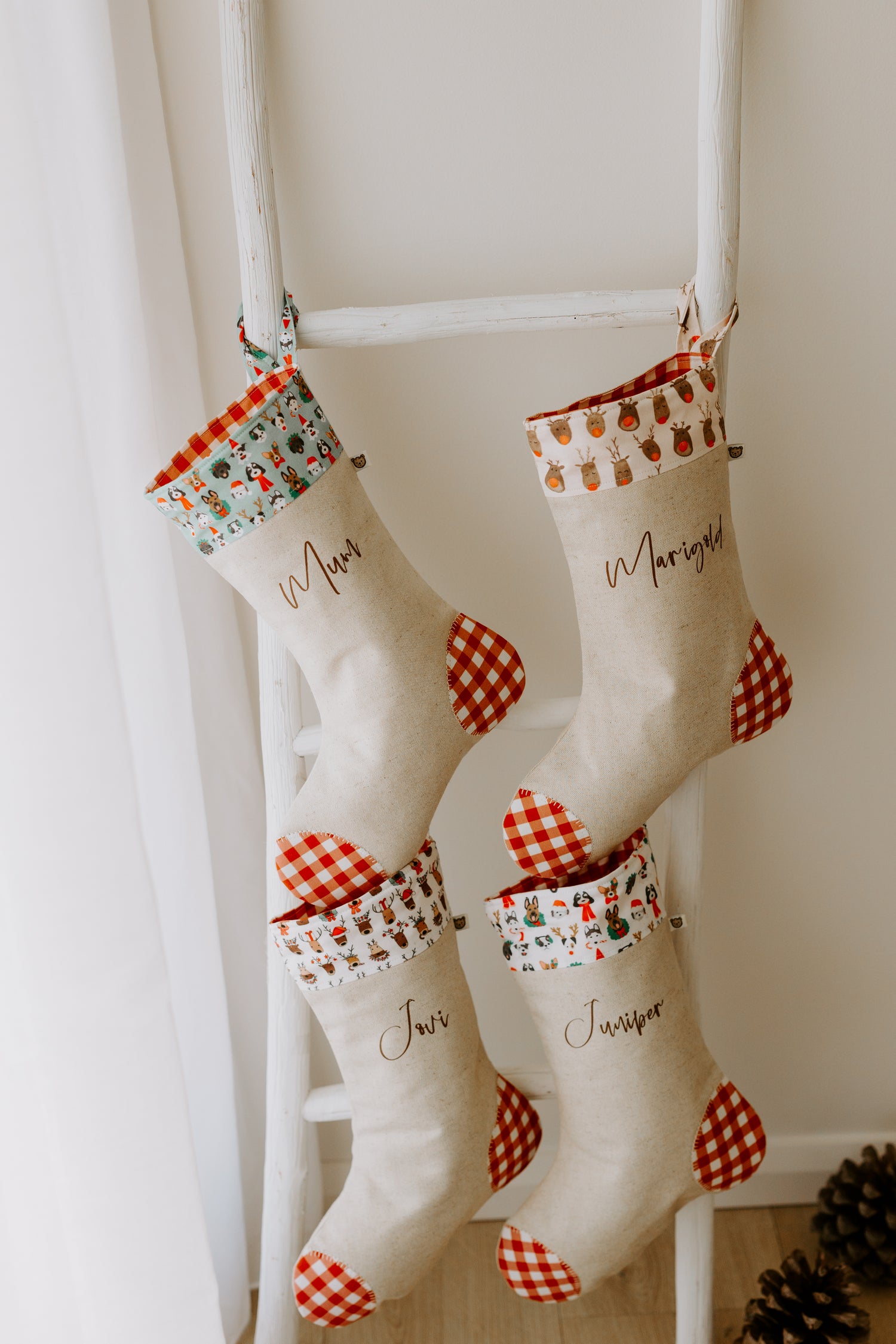 Set of four handmade personalised Christmas stockings hanging on a rustic wooden ladder, crafted in natural linen with bold red gingham heels and toes. Each stocking has a unique festive cuff design including Christmas puppies, reindeer, and holiday motifs, with names such as “Mum,” “Marigold,” “Jovi,” and “Juniper” in brown script. Perfect as custom family stockings, children’s Christmas gifts, or rustic holiday décor for Australian homes.