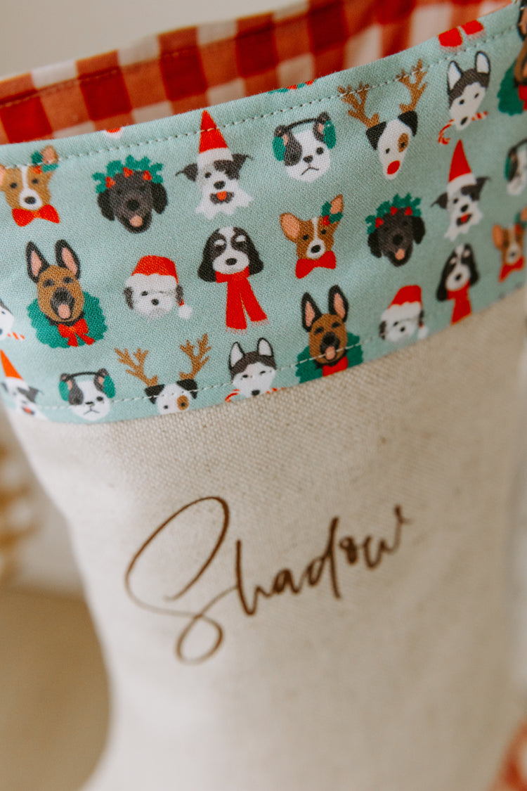 Personalised Christmas stocking with “Shadow” written in script font, featuring a festive cuff patterned with different dog breeds dressed in holiday outfits. The fabric shows huskies, spaniels, shepherds, and terriers wearing Santa hats, scarves, reindeer antlers, and Christmas wreaths. The natural linen stocking with red gingham trim is designed as a special pet stocking for dogs, perfect for fur babies and animal lovers in Australia.