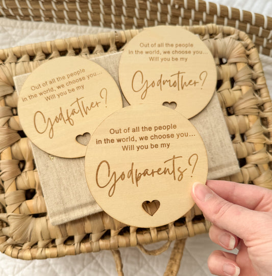 Three wooden tags asking if you will be godparents, held over a woven basket.