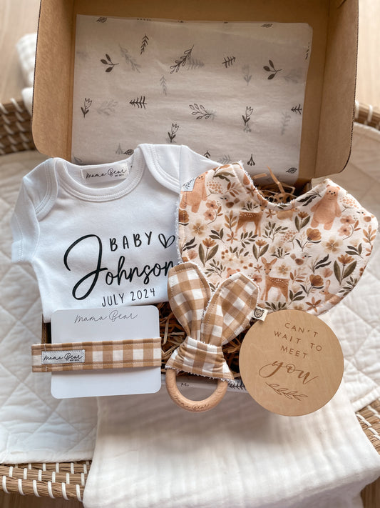 Personalised Baby Announcement Gift Set displayed in a Kraft brown gift box, including a white baby onesie personalised with ‘BABY Johnson July 2024’, handmade woodland bib with illustrations of a deer , bear, bunny , earthy tone leaves and flowers, a round milestone disc that says ‘can’t wait to meet you’ , teething ring with caramel check fabric on it and a matching dummy clip. 