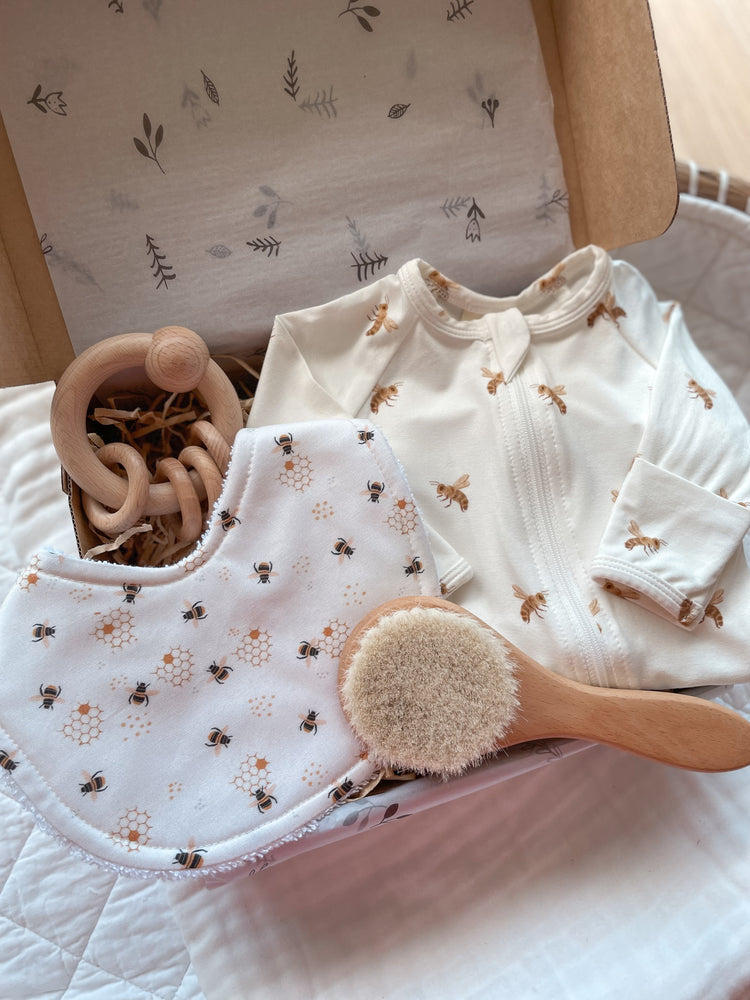 Gender neutral baby gift box featuring handmade bib and blossom & pear zipsuit with honey bee pattern on white background. Presented in a Kraft gift box filled with tissue paper and raffia. Also shown in the set is a wooden round baby hairbrush and a round wooden ring rattle.