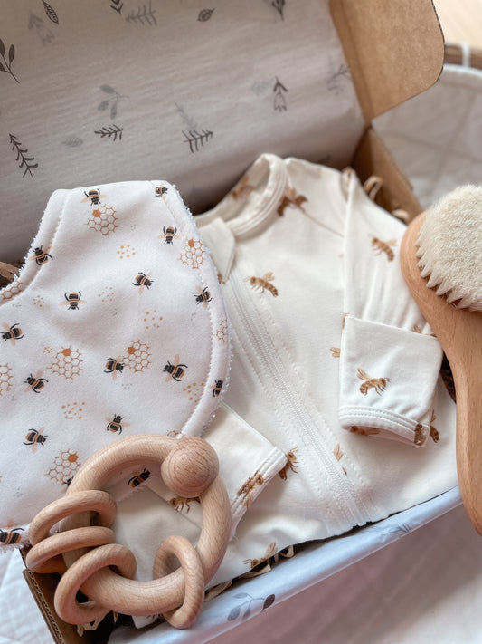 Gender neutral baby gift box featuring handmade bib and blossom & pear zipsuit with honey bee pattern on white background. Presented in a Kraft gift box filled with tissue paper and raffia. Also shown in the set is a wooden round baby hairbrush and a round wooden ring rattle.