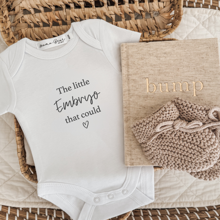 White baby announcement bodysuit that says "The little Embryo that could" with a small heart in black text. Flay lay in a woven basket with a linen bump journal and beige baby booties