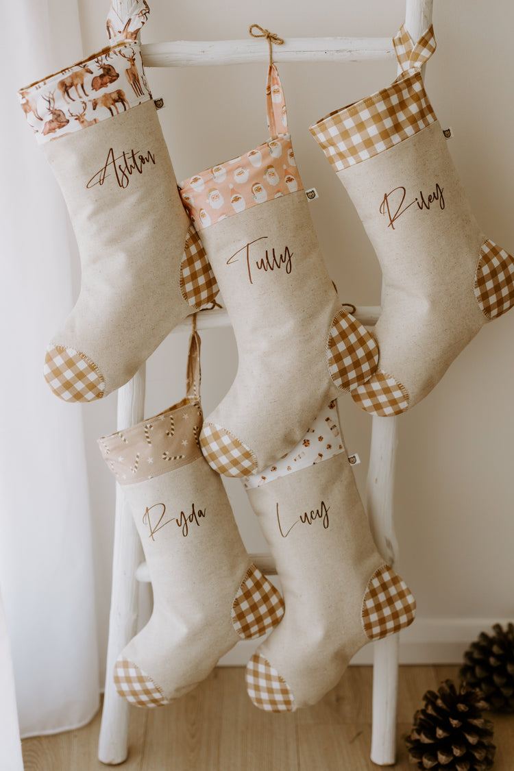 Set of handmade personalised Christmas stockings in beige fabric with caramel gingham heel and toe, featuring assorted festive cuffs including reindeer, Santa, candy cane and floral prints, customised with names Ashton, Tully, Riley, Ryda and Lucy.