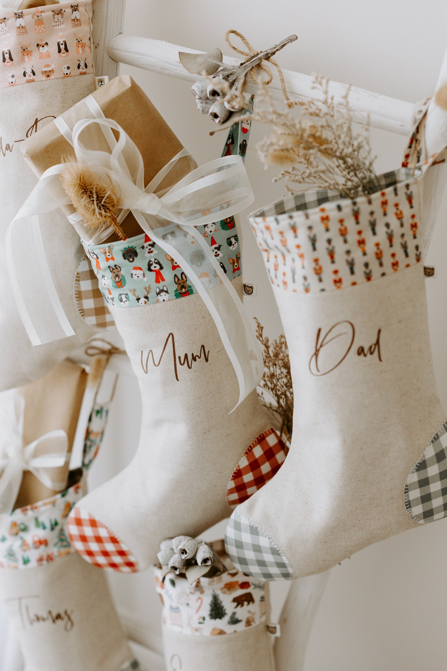 Handmade personalised Christmas stockings in beige fabric with red and green gingham detail, featuring festive dog print cuff for Mum and nutcracker print cuff for Dad, filled with rustic holiday décor.