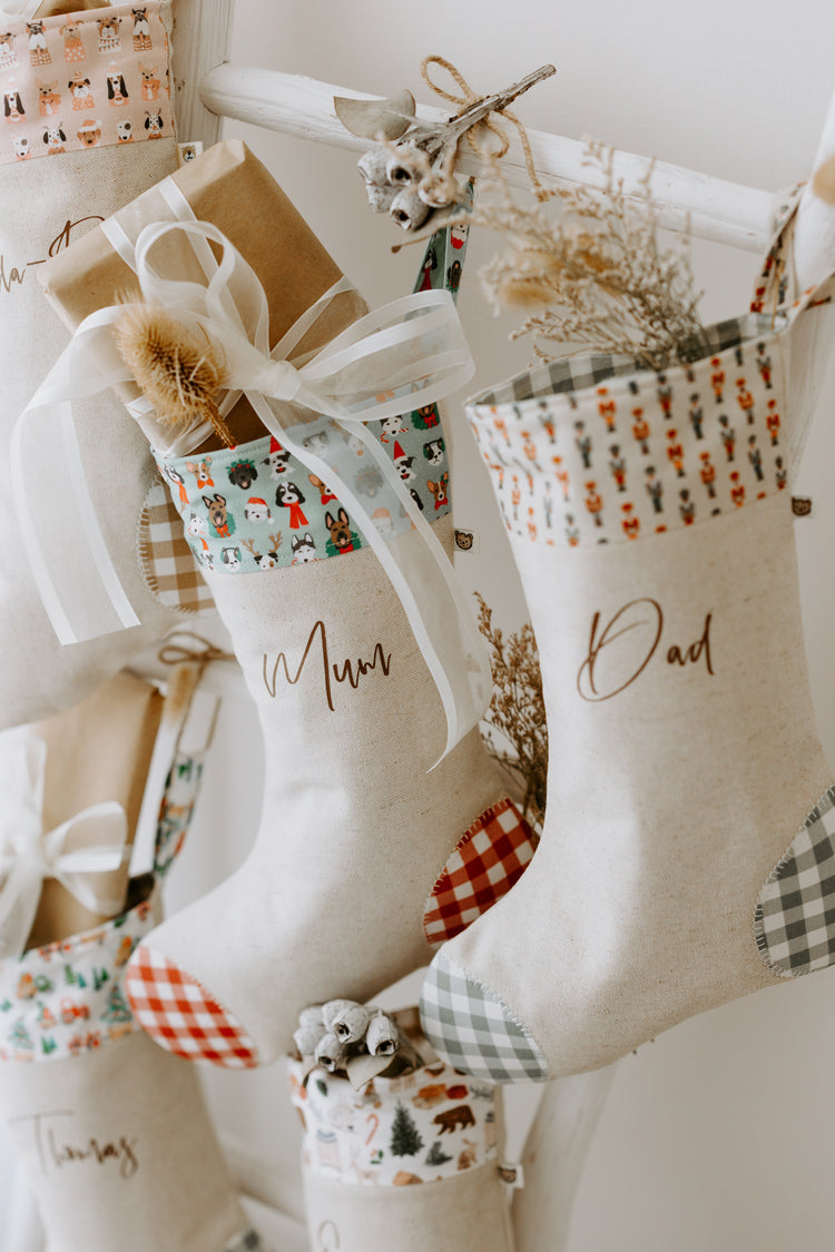 Handmade personalised Christmas stockings in beige fabric with red and green gingham detail, featuring festive dog print cuff for Mum and nutcracker print cuff for Dad, filled with rustic holiday décor.