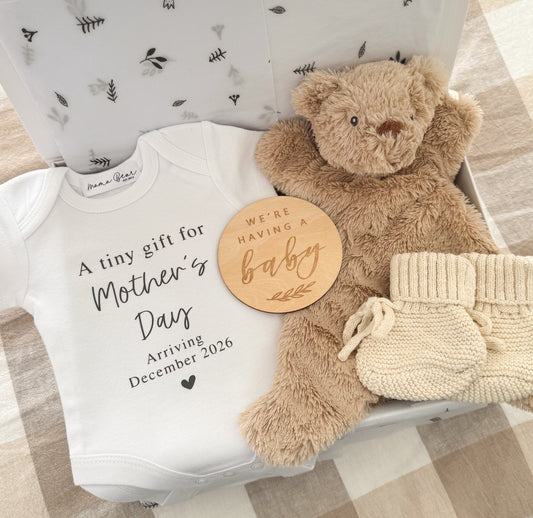 Baby announcement onesie for Mother's Day saying 'A tiny gift for Mother's Day arriving December 2026'. With a soft brown teddy bear comforter and wooden disc saying 'We're having a baby' and some honey baby booties packaged inside a white gift box.