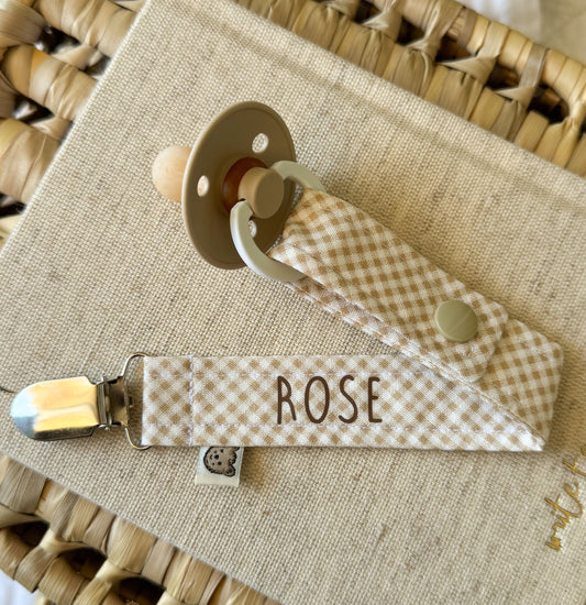 A baby dummy clip in a soft beige and cream mini gingham print. Personalised in brown 'ROSE'.