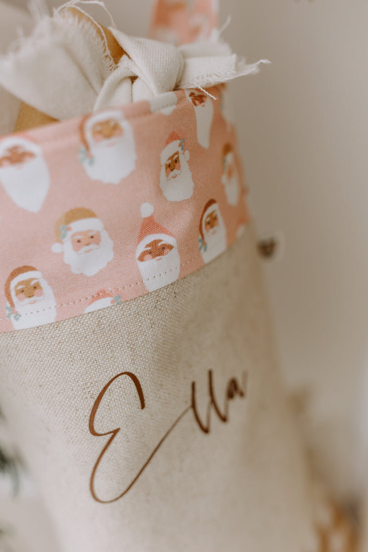 Custom name Christmas stocking with whimsical pastel pink Santa face design fabric cuff, script font personalisation, and gingham accents. Perfect gift idea for a baby girl